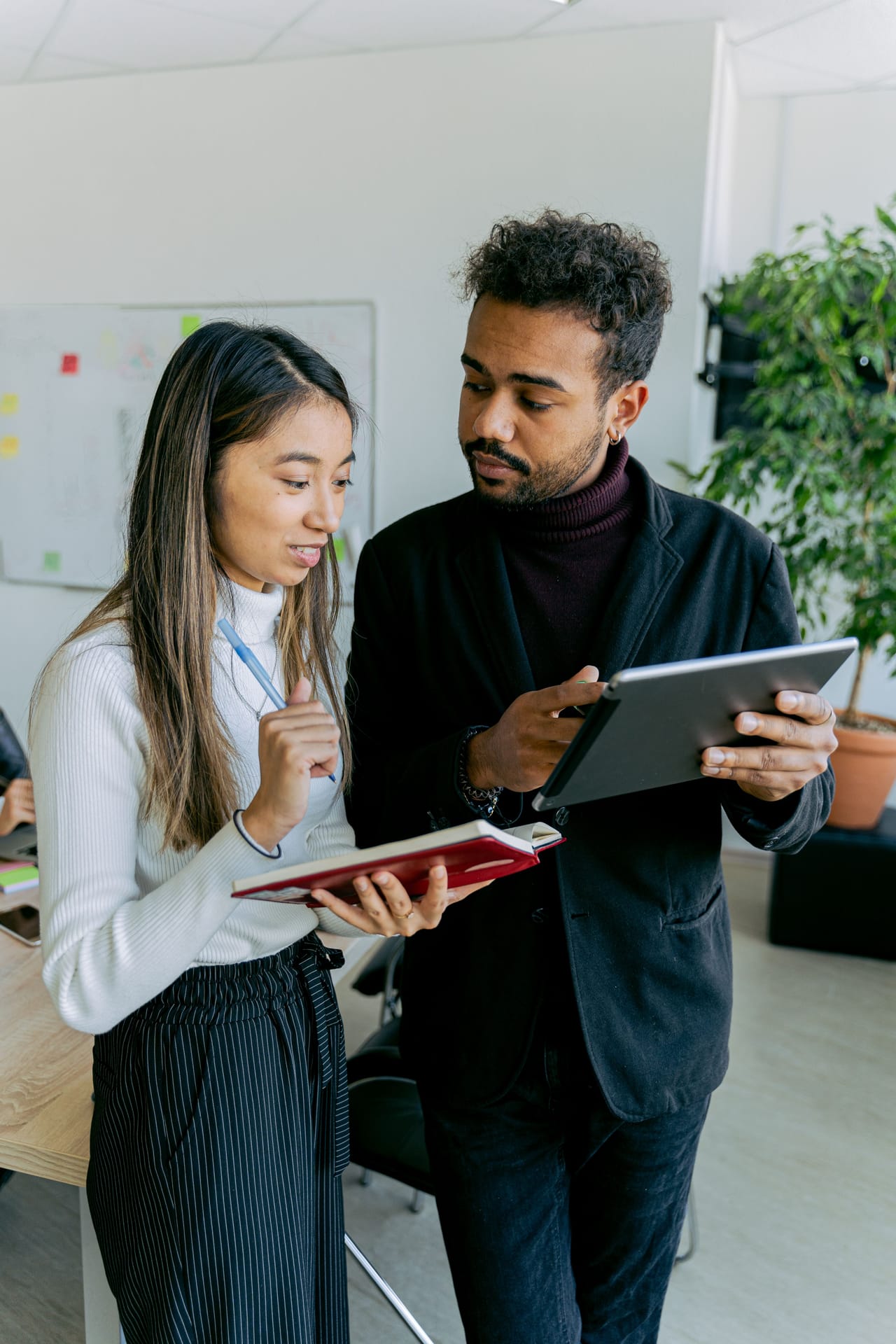 Two colleagues standing together in an office, one holding a notebook and the other a tablet